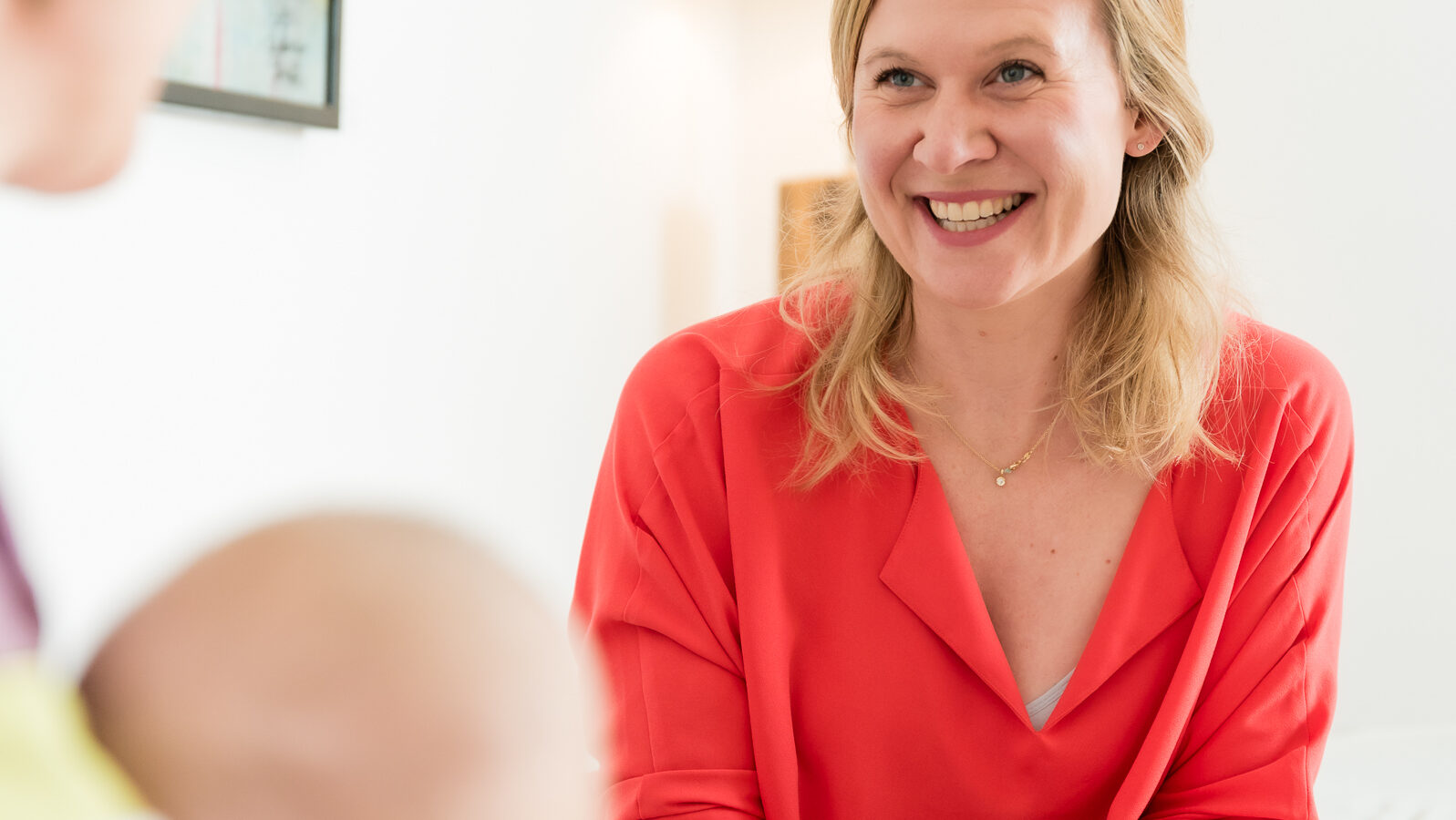 A woman in a red shirt smiles while sitting at a table. She is facing another person who is holding a baby, partially visible. A lamp and a painting are in the softly lit background.