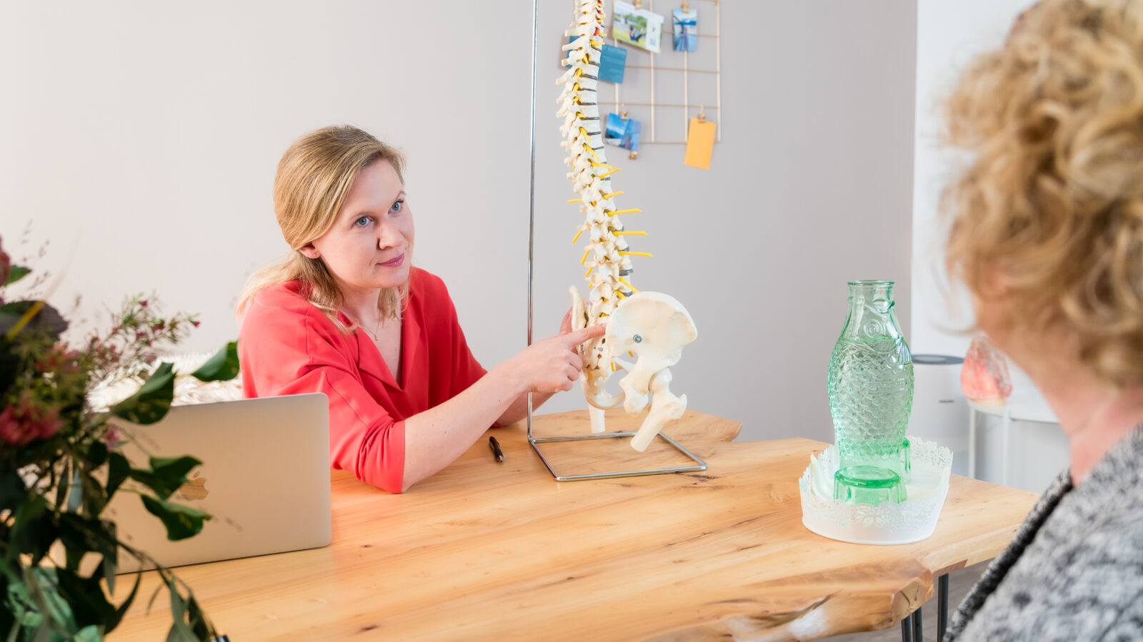 A woman, seated at a wooden table in an office, explains a spine model to a person sitting across from her. A laptop, a large glass jug, a vase with flowers, and various notes and items adorn the table and walls.