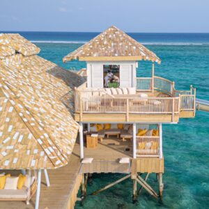 A luxurious overwater bungalow with thatched roofs extends over a clear blue ocean at a Maldives luxury private resort. The villa features open-air living spaces with seating areas and a water slide that leads directly into the sea. A person can be seen relaxing on a lounger in the distance.