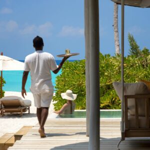 A person in white attire carries a tray while walking on a wooden deck toward a woman with a large sun hat sitting by a private pool at a Maldives luxury resort. In the background, there are lounge chairs with umbrellas, lush green foliage, and a turquoise ocean under a clear blue sky.