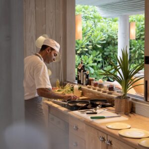 A chef in a white uniform and hat is cooking at a well-organized kitchen counter in one of the Maldives Luxury Private Resort Villas. Various ingredients and utensils are spread out, and leafy plants outside the window provide a lush, green backdrop. The kitchen has a warm and inviting ambiance.