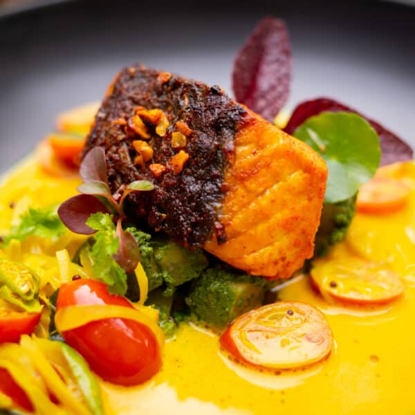 A Thailand Exclusive Experience featuring a seared piece of fish, garnished with herbs and spices, atop a vibrant yellow sauce. Accompanied by colorful vegetables, including cherry tomatoes and greens, all arranged on a black plate at Soneva’s signature restaurant.