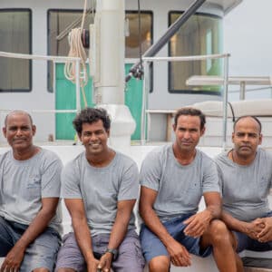 Four men dressed in grey t-shirts and navy shorts are sitting on the steps of a boat. The background shows parts of the boat, including railings, a mast, and a seating area. They all have a friendly demeanor and are posing for the camera, embodying the team spirit of their Soneva careers.