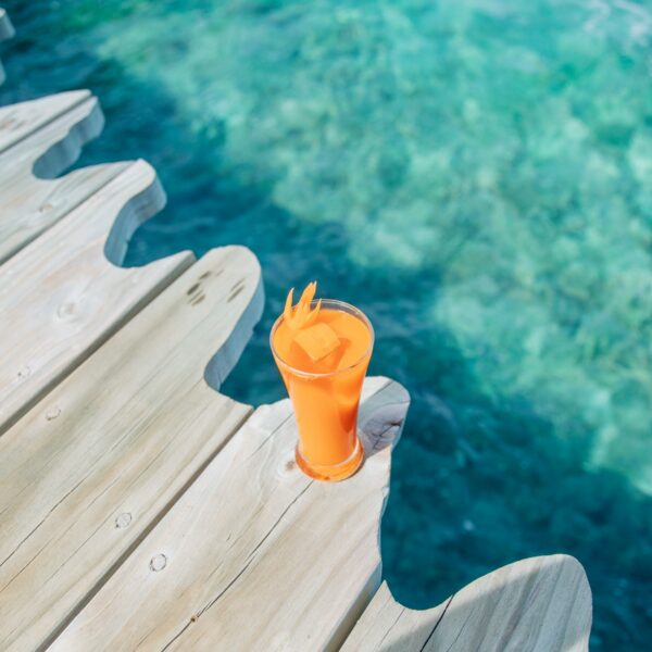 A refreshing orange drink in a tall glass sits on a wooden deck with a clear blue ocean in the background. The deck has a unique wavy edge, and the glass has a straw and ice cubes. The vibrant turquoise water showcases Soneva Exclusive Offers, blending natural beauty with luxury experiences.