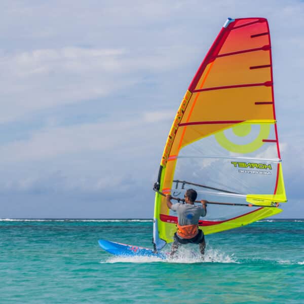 A windsurfer glides across turquoise waters on a sunny day during an exclusive experience in the Maldives. The colorful sail, predominantly red and yellow with a large green circle, stands out against the blue sea and sky. The person is wearing a gray shirt and orange life vest, pushing forward with the wind.