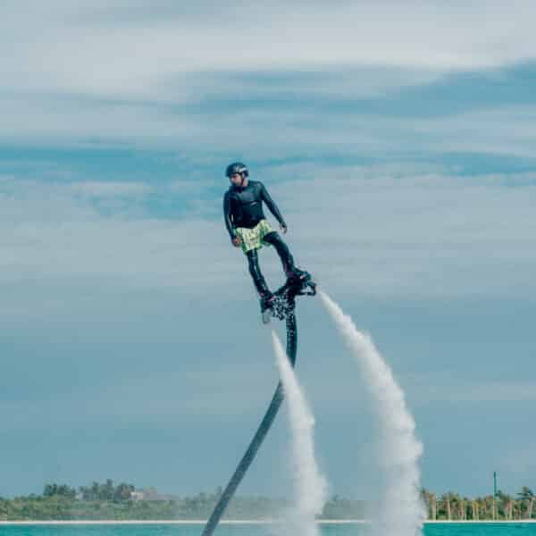 A person wearing a black helmet and safety gear is flyboarding over the ocean. Water jets propel them high above the water's surface, with clear skies and distant land visible in the background, creating a Maldives exclusive experience at Soneva.