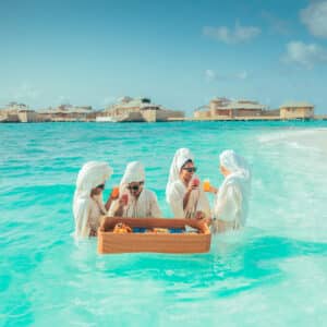 Four people, possibly on their Maldives honeymoon, are waist-deep in turquoise water near a sandy shore, enjoying drinks and snacks from a floating wicker basket. They are wearing white outfits and head coverings, with overwater bungalows visible in the background under a partly cloudy sky.