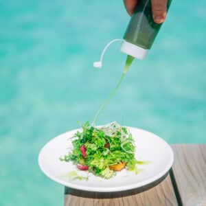 A hand pours green dressing onto a fresh salad on a white plate. The plate is placed on a wooden surface with a turquoise water background, creating a vibrant and refreshing scene—much like the paradisiacal settings of Soneva Exclusive Offers.