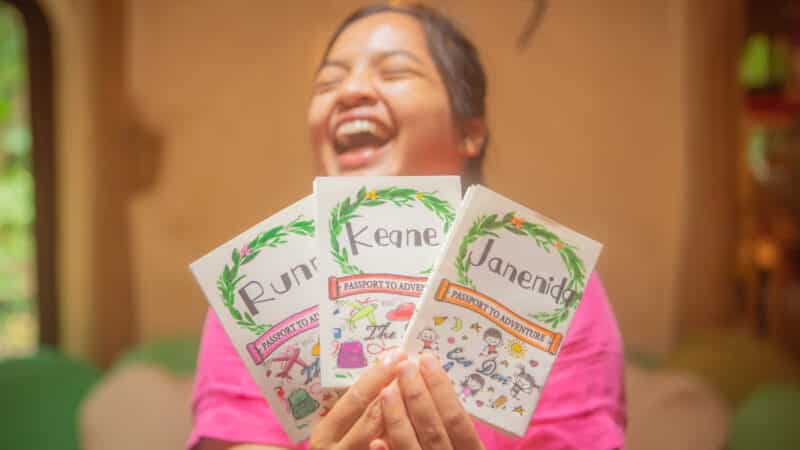 A person wearing a pink shirt is joyfully holding up three passports labeled "Keane," "Runa," and "Janenia." The passports, part of a Thailand Exclusive Experience at Soneva, have festive, colorful designs and illustrations. The background appears to be a warmly lit indoor space.