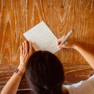 A person with dark hair, dressed in a white shirt, writes a handwritten letter on a piece of paper with a pen. Viewed from above, the scene reveals a wooden table. One hand holds the paper steady while the other writes what seems to be plans for their upcoming Maldives honeymoon after their wedding.