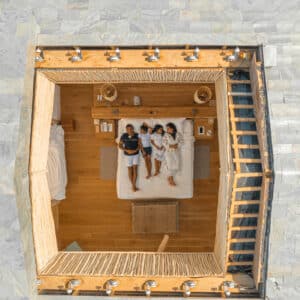 A top-down view of a modern, open-roof bedroom with a double bed. Two people are lying side by side on the bed, reading or using a tablet. The room features wooden flooring and minimalistic decor, evoking the luxury of Soneva Exclusive Offers, with an attached staircase leading up to the roof.