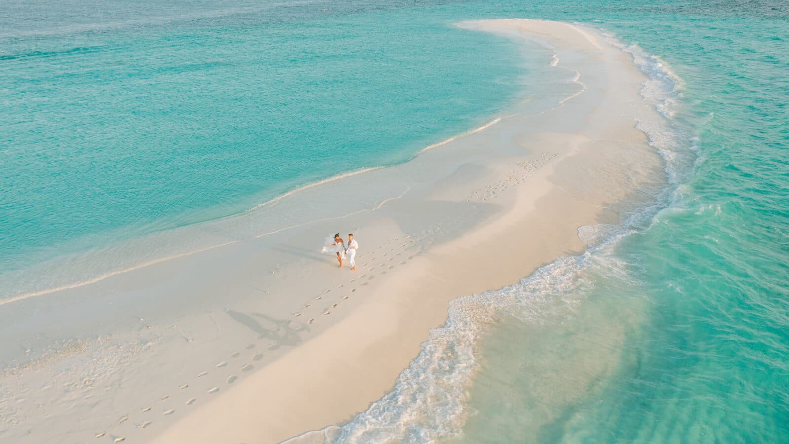 An aerial view of a couple walking hand in hand along a narrow, curved sandbar surrounded by the turquoise ocean of the Maldives. Their footprints trail behind them, and gentle waves lap at the sandy shore. The scene is serene and idyllic, epitomizing a tropical paradise perfect for a honeymoon.