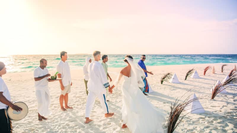 A bride and groom walk hand in hand on a beach, surrounded by wedding guests dressed in white. Palm fronds and white flags decorate the sand. The ocean waves roll in the background under a clear sky as musicians play traditional drums, creating a perfect scene reminiscent of Soneva Exclusive Offers.