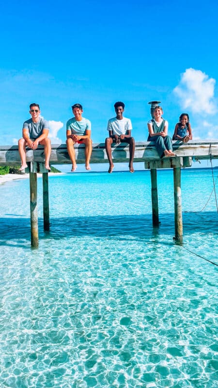 Five people sit on a wooden bridge above clear turquoise water, with a sandy beach in the background. They appear to be enjoying a sunny family holiday at the Soneva resort in the Maldives, all facing forward and relaxed. Two men are on the left, followed by two younger boys and a little girl on the right.