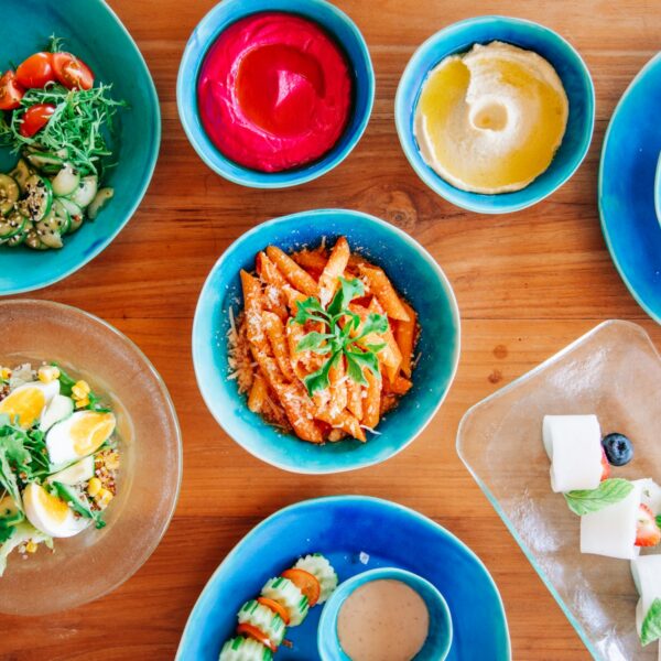 A colorful spread of dishes on a wooden table includes grilled vegetables on skewers, vibrant beetroot dip, hummus, a mixed grain salad, a fresh salad with eggs, penne pasta in tomato sauce, and a dessert platter with pieces of cheese and blueberries—perfect for a family holiday reminiscent of dining at Soneva in the Maldives.