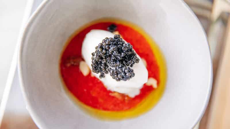 A close-up of a gourmet dish featuring a dollop of white cream topped with black caviar, placed on a vibrant red sauce, served in a light grey bowl. The elegant presentation evokes the sophistication one would expect at a Maldives luxury resort like Soneva.