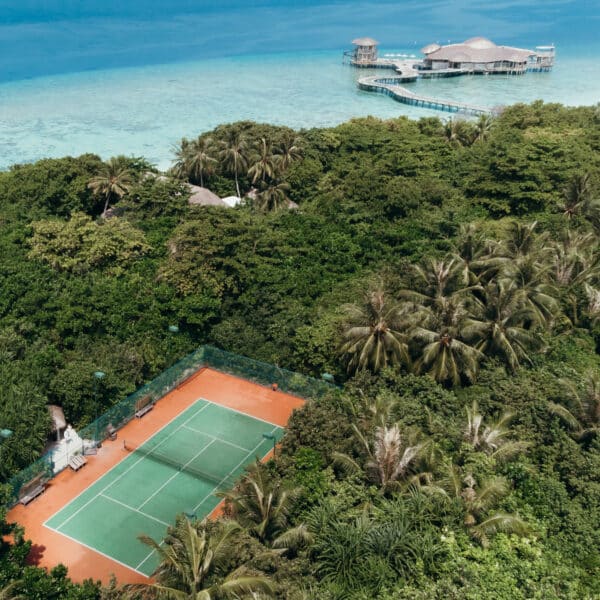 Aerial view of a red clay tennis court surrounded by dense green foliage and palm trees, with a path leading to a wooden pavilion over clear blue water in the background. This exclusive experience on a tropical island embodies the serene beauty Soneva is renowned for, reminiscent of the Maldives.