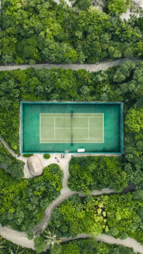 Aerial view of a tennis court surrounded by dense, lush green forest. Pathways meander through the trees, leading to the court, which stands out with its vibrant green surface. A small hut is visible near the court's lower left corner, reminiscent of a Maldives exclusive experience at Soneva resorts.