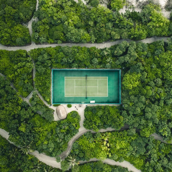 Aerial view of a tennis court surrounded by dense, lush green forest. Pathways meander through the trees, leading to the court, which stands out with its vibrant green surface. A small hut is visible near the court's lower left corner, reminiscent of a Maldives exclusive experience at Soneva resorts.