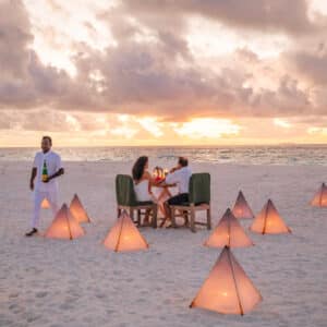 A couple sits at a dining table on a beach, surrounded by illuminated triangular lanterns. A waiter, dressed in white, approaches the table with a bottle in hand. The sky is filled with dramatic clouds as the sun sets over the ocean in the background—a perfect setting for a Soneva Career experience.