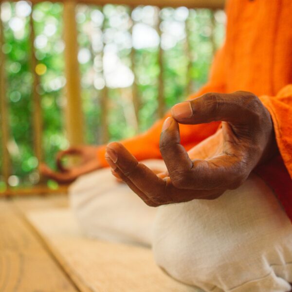 Person in vibrant orange clothing sits cross-legged on a wooden floor with their hands on their knees in a meditative pose, embodying a Thailand exclusive experience. The image focuses on the hand making a mudra gesture, with a blurred natural background visible, reminiscent of Soneva&#039;s serene retreats.