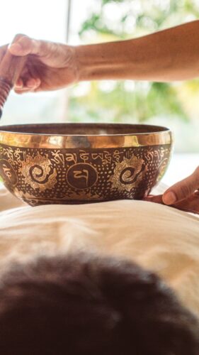 A person holding a traditional Tibetan singing bowl over another person lying on their back with a white sheet covering them, set in an outdoor environment with greenery in the background – a Thailand exclusive experience brought to you by Soneva.