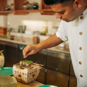 A chef garnishing skewered dishes with herbs in a restaurant kitchen. The skewers are placed on a small grill atop wooden containers with Japanese characters. The countertop in the background, adorned with various kitchen items and utensils, reflects the precision and elegance of a Soneva Career.