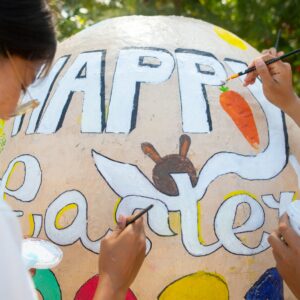 Three people are painting a large Easter egg sculpture outdoors. The egg features the words "Happy Easter" along with a bunny and carrot design. One person, smiling and wearing bunny ears, enjoys this festive family holiday activity. Trees form the background, indicating a sunny day reminiscent of Maldive Soneva's serene surroundings.