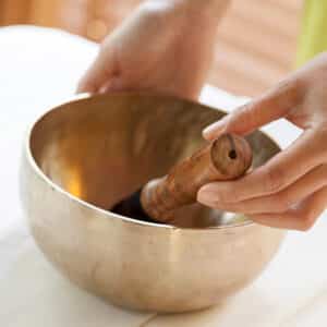 Close-up of a person holding a Tibetan singing bowl, with both hands on either side of the bowl and a wooden mallet resting inside. The setting appears calm and serene, suggesting a meditative or healing atmosphere, reminiscent of Soneva Exclusive Offers&#039; tranquil retreats.