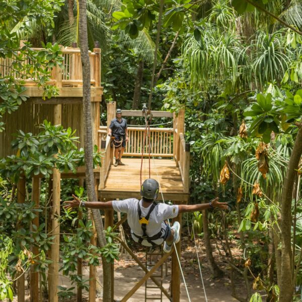 A person wearing a harness is seen zip-lining through a lush, green tropical forest towards a wooden platform where another stands. The dense foliage and interconnected treehouses evoke the thrill of a Maldives exclusive experience at Soneva, creating an unforgettable outdoor adventure setting.