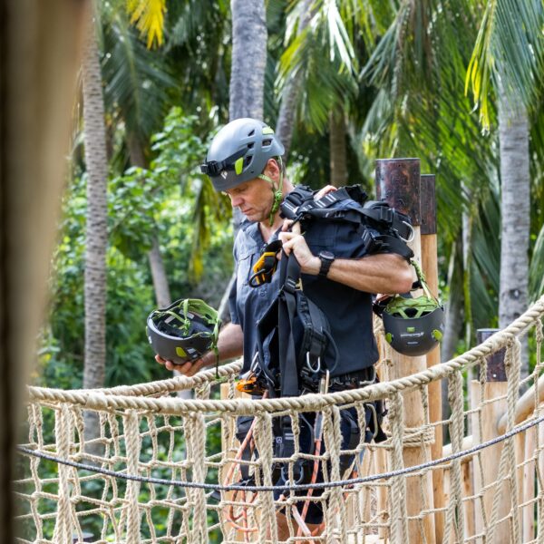 A person wearing a black helmet and harness walks across a rope bridge in a tropical forest. They hold another helmet and additional gear, making their way through the lush greenery of this Maldives exclusive experience at Soneva. Palm trees sway gently in the background.