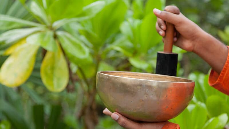 A person in an orange sleeve holds a bronze singing bowl in one hand and a wooden mallet in the other, embodying the tranquility of a Maldives spa. The background is lush with green leaves, suggesting an outdoor, nature-filled setting perfect for wellness and relaxation.