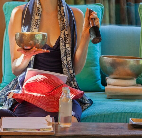 A person sits on a turquoise couch holding a singing bowl in one hand and a mallet in the other, embracing a Thailand exclusive experience. Another singing bowl rests beside them, with a small glass bottle and red cloth on their lap. A piece of paper lies on the wooden table in front of them.