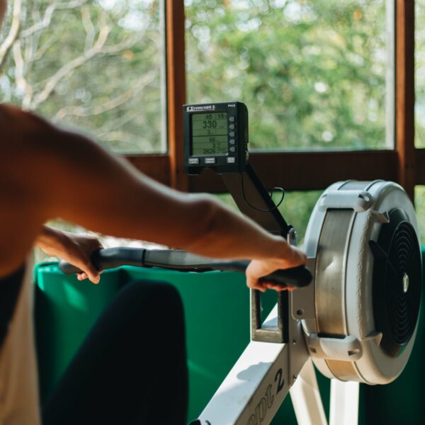 A person in athletic wear uses a rowing machine indoors, with a window view of green trees outside. The machine&#039;s display screen shows workout stats. This Thailand exclusive experience suggests an exercise routine in a home or gym setting with natural light coming through the window.