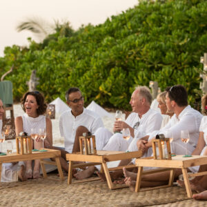 A group of people dressed in white outfits sit around low wooden tables on a beach at Soneva Fushi. They are smiling and engaging in conversation, surrounded by lush greenery. Some have drinks in their hands, and the tables hold candles and wine glasses, enjoying a quintessential Maldives dining experience.