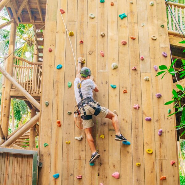 A person wearing a helmet and harness climbs a wooden rock climbing wall with colorful holds at the Soneva outdoor adventure park. Surrounded by lush greenery and palm trees, this Maldives exclusive experience captures an exhilarating mid-climb moment.