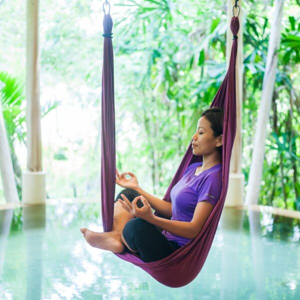 A woman sits in a maroon aerial yoga hammock, meditating with her legs crossed and hands in a mudra pose. She is wearing a purple shirt and black pants. The lush, green backdrop features tropical plants and trees, with a tranquil pool below—a Thailand exclusive experience at Soneva.