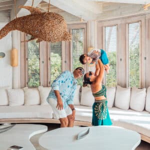 A family of three enjoys a moment in a sunlit room with large windows, reminiscent of a tranquil Maldive Soneva retreat. The father, in a blue shirt and white shorts, smiles as the mother, wearing a green dress, playfully lifts their young child. A decorative woven dolphin hangs from the ceiling.