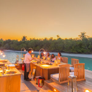 A group of people enjoys a meal at a beachfront wooden bar under a golden sunset sky, dining at Soneva Jani. A waiter in a white shirt serves them. Surrounded by calm water and lush greenery, the setting exudes a tropical, serene vibe.