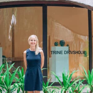 A person with long blonde hair, wearing a sleeveless black dress, is standing outdoors in front of a building. Behind them, a glass window displays &quot;TRINE DRIVSHOLM.&quot; Lush green plants are in the foreground, creating a scene that evokes the exclusivity of a Maldives experience like Soneva.