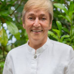 A smiling older woman with short, light-colored hair stands in front of a background of lush, green foliage. She is wearing a white, button-up shirt, embodying the vibrant spirit of a Soneva Career.