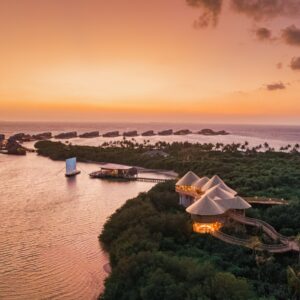 A tranquil island resort at sunset features overwater bungalows connected to the main island via walkways. Tropical foliage covers the island, a sailboat glides on calm water, and the sky is painted in warm orange and pink hues, while guests indulge in Maldives spa wellness experiences.