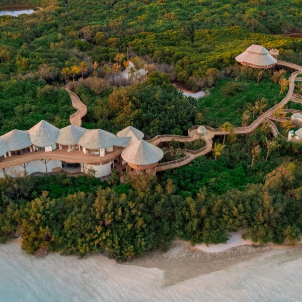 Aerial view of a luxurious resort surrounded by dense greenery and near a sandy shoreline. The resort, resembling a Maldives spa, features several thatched-roof structures connected by elevated wooden walkways, blending harmoniously with the lush foliage and natural landscape to offer ultimate wellness.