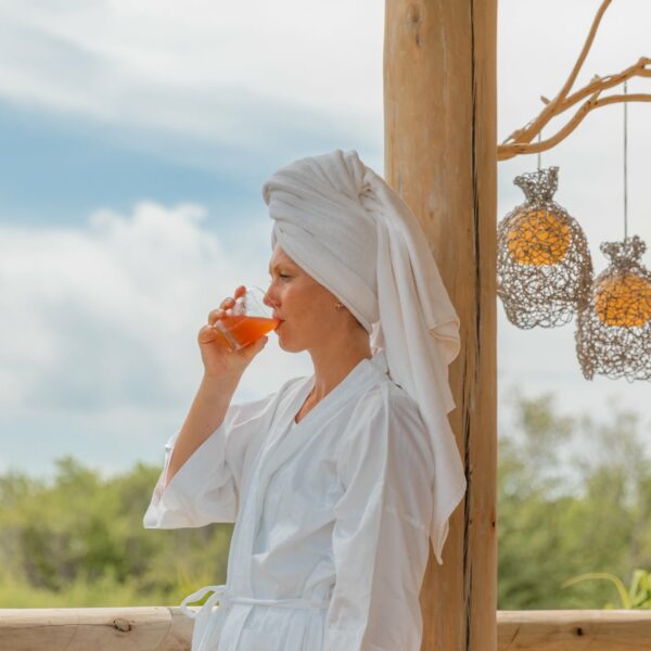 A person wearing a white robe and a towel wrapped around their head stands on a wooden deck, sipping a drink. Decorative lanterns hang from a tree branch beside them, evoking the ambiance of Soneva Exclusive Offers. The background features lush greenery and a partly cloudy sky.