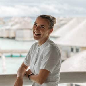 A woman with blond hair tied back is leaning against a wooden railing, smiling broadly. She is wearing a white shirt. The background shows an overwater bungalow resort with a cloudy sky above, perhaps hinting at her fulfilling Soneva Career in this picturesque paradise.