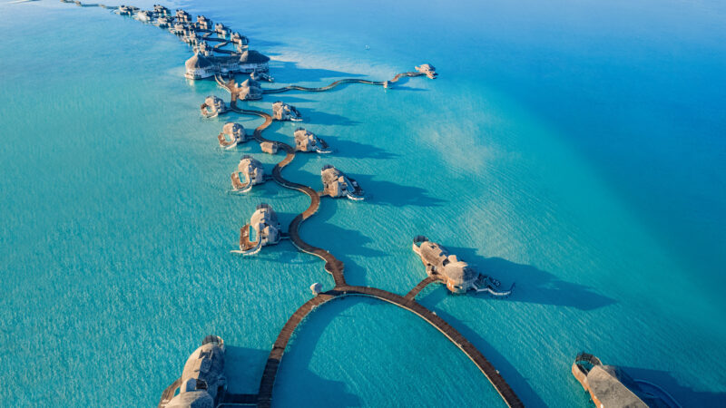 Aerial view of luxurious overwater bungalows connected by wooden walkways in the turquoise waters of a tropical island resort. The arrangement forms a curved shape, extending from the shoreline into the clear blue sea. Lush greenery is visible on the far left—a perfect Soneva Maldives holiday.