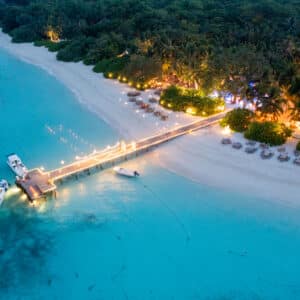 Aerial view of a picturesque tropical beach with turquoise water at Soneva in the Maldives. A long, illuminated pier extends into the sea, where boats are docked. The sandy shore is lined with palm trees, loungers, and small, warmly lit structures—ideal for a family holiday amid dense greenery.