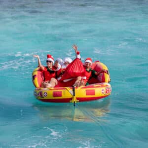 A family of four, donning Santa hats, sits in a bright yellow and red inflatable raft, waving and smiling excitedly as they float on the clear turquoise waters of the Maldives. The raft, tethered with a rope, drifts gently near Soneva. One person holds a large red sack brimming with holiday cheer.