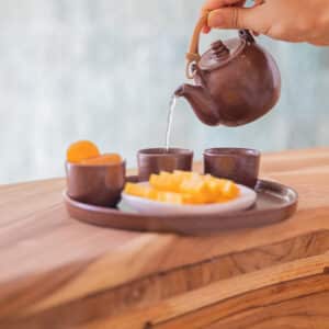 A hand is pouring tea from a small brown teapot into a matching teacup. The teacup sits on a wooden tray with two other small teacups and a bowl of sliced fruit, evoking the tranquility of a Maldives spa experience. The background is softly blurred, enhancing the scene's sense of wellness.