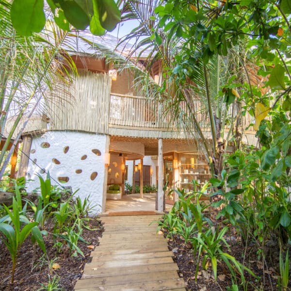 A wooden pathway leads to a rustic two-story building surrounded by lush greenery. The building features a mix of wooden and white plaster walls with circular cutouts, and has a balcony on the upper level, creating a tropical, eco-friendly atmosphere reminiscent of an exclusive Maldives experience at Soneva.